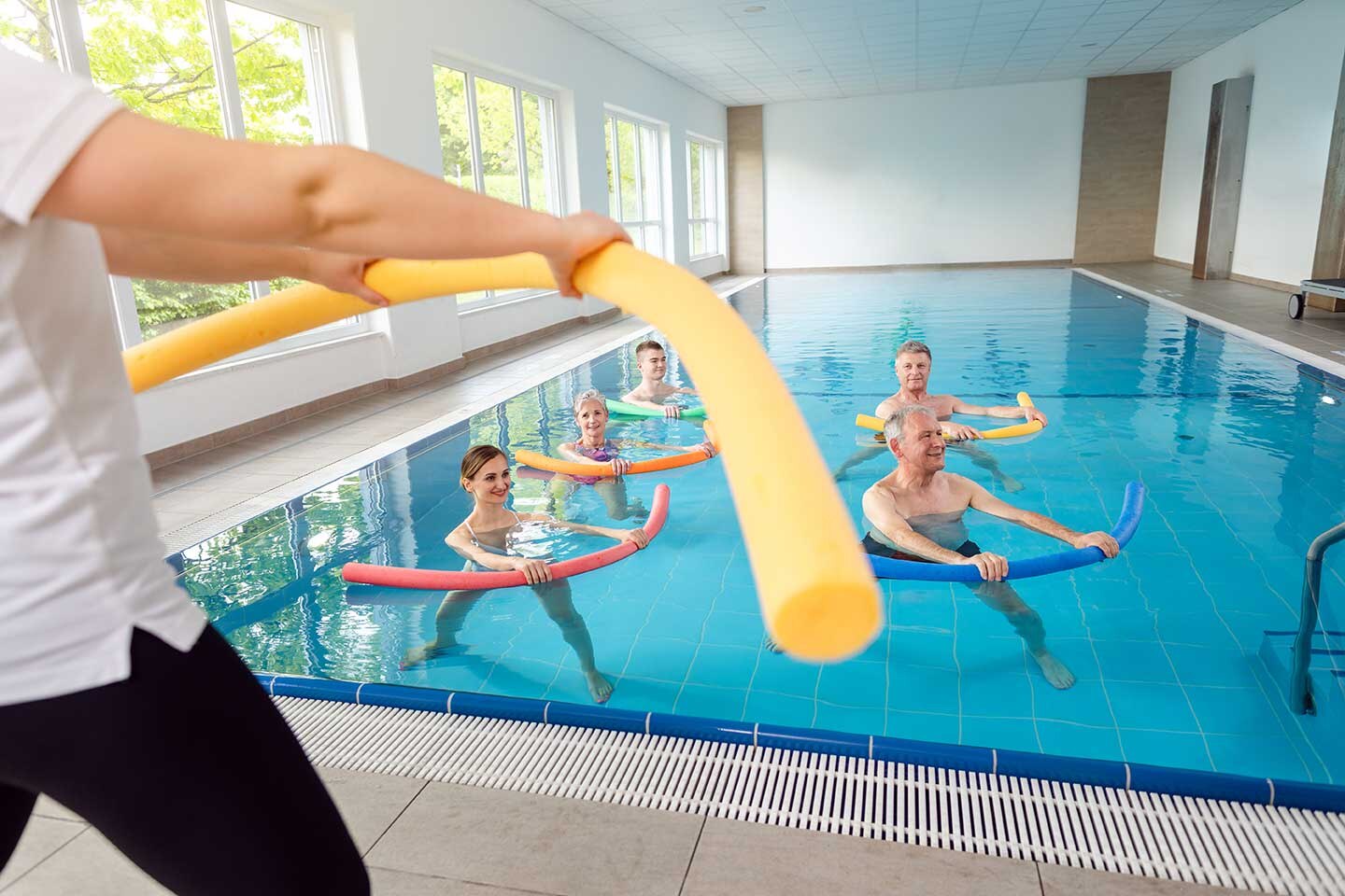 Ein Bademeister steht vor einem Pool mit Menschen, die Aquafitness betreiben. Sie haben Poolnudeln in der Hand.